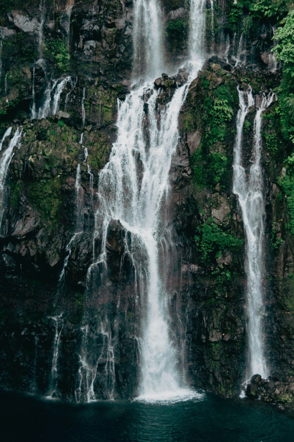 Several waterfalls cascading down a dark, mossy volcanic cliff into a deep pool, Reunion Island